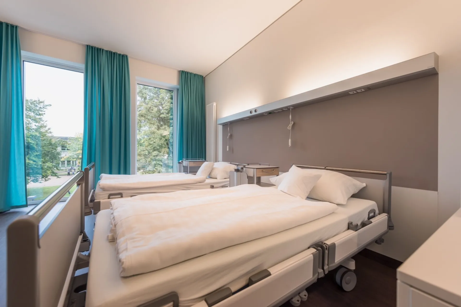 Two side-by-side hospital beds with white duvets in a bright room with large windows and green curtains